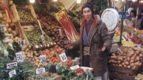 A stallholder wearing a brown coat and blue hat gestures next to her vegetable stall.