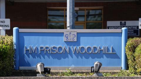 Blue sign with grey writing reading HM Prison Woodhill. There are lights at the bottom of the sign pointing upwards and hedges either side. 