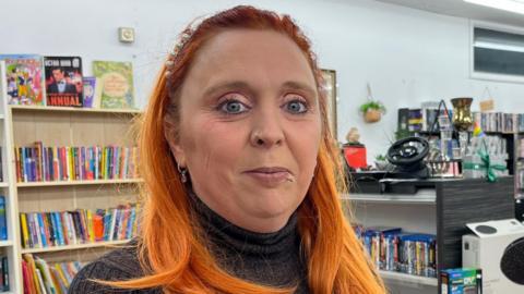 Lianne Simpson is stood in the middle of the image. She has long orange hair and is wearing a pearl headband and a grey turtle neck jumper. Behind her are rows of bookcases. 