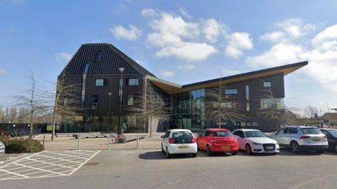 A modern-looking, dark coloured building with a sloping roof and cars parked outside. 