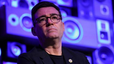 Andy Burnham looks serious while standing in front of a blue and purple background.