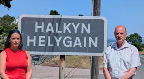 A woman in a red top and brown hair and a balding man in a pale blue shirt. They're stood outside a village in front of a sign that says 'Halkyn Helygain' .