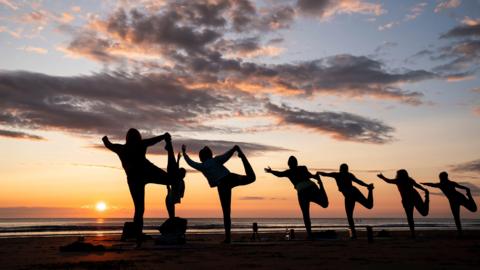 Six silhouettes in a line holding a yoga pose consisting of them holding their left hand out in front of them and their right leg behind them, a teacher is in front of them. During sunset on the beach with a few clouds in the sky.