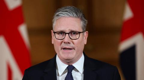 Keir Starmer. He has short grey-ish hair, wearing glasses, white shirt and dark blazer and tie. Two British flags are in the background.