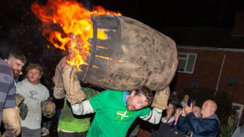 A man in a green t-shirt carrying a barrel that is on fire as people watch on