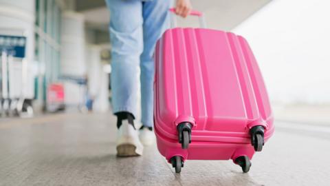 A young woman wearing light jeans and white trainers pulling a bright pink suitcase in an airport