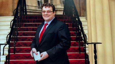 David Sparkes wearing a suit and holding an award. There are red-carpeted stairs behind him and a long, beige curtain.