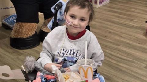 Young girl, 8, sat in a chair with a smile on her face looking straight ahead. She's eating a fast food meal inside a hospital ward.