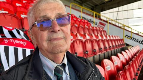 A man with short hair wearing glasses, dark jacket, a shirt and tie. He is sitting in a football stand in front of rows of folded red seats. Behind him is a black and white striped Grimsby Town shirt. On the shirt is the logo of the club sponsor, Myenergi, written in white letters on a red background.