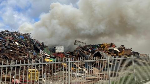 A plume of smoke billowing from a scrap yard. The grey smoke is set against a blue sky.