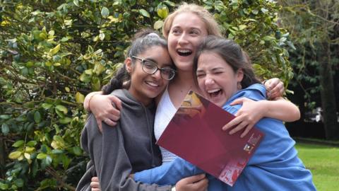 Three students are hugging. One is holding a school folder.