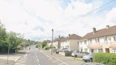 A suburban road with semi-detached houses on one side. Telegraph poles, parked cars and other street furniture is visible.