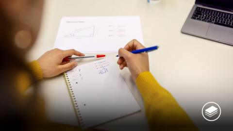 Woman sitting at desk and taking maths notes with a pen. A laptop is in the corner. 