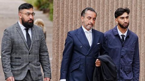 A man in a double breasted suit jacket walks along with a young man on either side of him. The one on the left wears a grey checked suit and the one on the other side wears a navy blazer. 