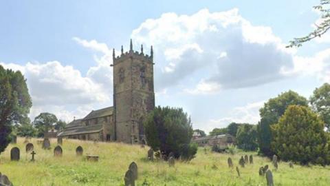 The 10th century St Peter's Church in Felkirk with a traditional view of a grassy churchyard, gravestones, a yew tree and other trees, and a stone wall