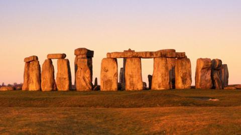 Image of Stonhenge at dusk. Large rectangular stones stand in a circle, some with other stone laid on top to bridge the gap between the stones. The surrounding land is covered in green grass and the horizon is orange.