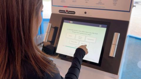You can see the side of an anonymous woman's face with long dark hair sitting in front of a machine with a screen, which she is tapping with her finger. 