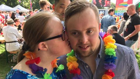 Michael Gallagher stands next to his girlfriend Alison Williams at a Pride event. They are both wearing colourful flower garlands around their necks and Alison kisses Michael's cheek. There are people behind them sitting on tables on grass and Pride banners.