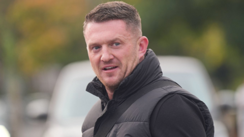 Political activist Tommy Robinson outside Folkestone Police Station. Robinson is wearing a black jumper and a black gillet. He is looking slightly away from the camera taking the image, has stubble and his hair gelled to one side.