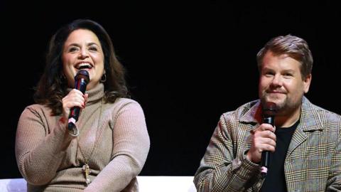 Ruth Jones on the left wearing a beige dress, and smiling. She has gold earrings and a long gold chain. She has dark hair and holds a mic to her hand. James Corden is on the right and has short dark-blonde hair and has a black t-shirt on under a checked jacket.