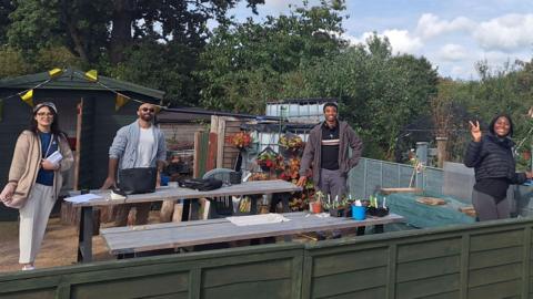 From left Kribina, Vishnu, Rotimi and Rosemary look at the camera and are smiling. They are stood next to a table with some paper work on and have a wooden shed and trees behind them. 