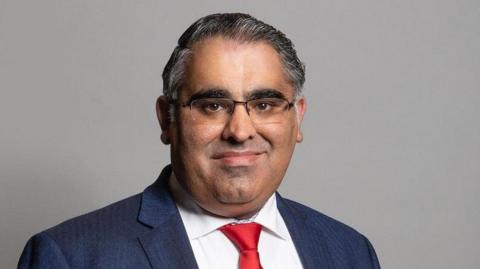 Tahir Ali pictured wearing a formal dark blue suit, white shirt and red tie