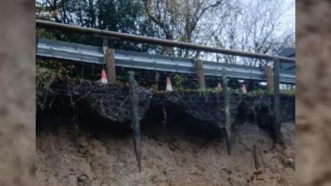 Close up shot of the side of the road with a grey fence in the foreground and trees in the background. There are orange cones by the fence too. Underneath the fence is exposed soil which shows signs of a large landslip.