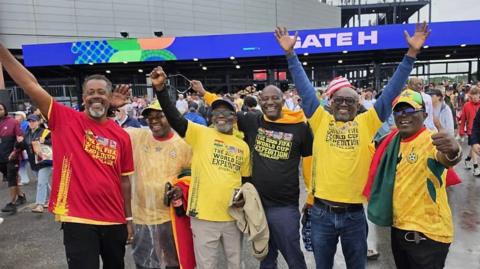 Six men stand in a line smiling and looking excited as they raise their arms in the air, wave at the camera and give a thumbs up. They are all wearing some combination of Ghana colours, predominantly yellow. Behind them, a crowd moves outside a stadium, with a blue video sign above an entrance which reads 'Gate H' in capital letters