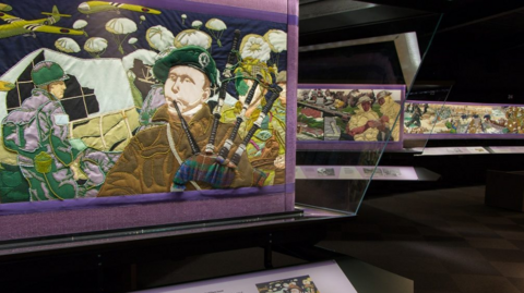 A general view of the Overlord Embroidery, with three sections pictured. In the one closest to the camera, a man is playing the bagpipes, with planes and 