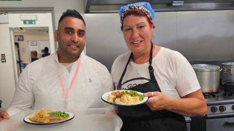 A man and woman stand in a kitchen, each is holding a plate of food. The man wears a plastic apron over his shirt, the woman wears a protective head covering.
