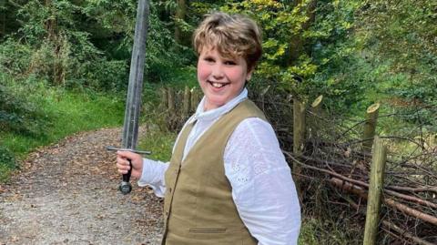 Wilf outside holding a sword and wearing a white shirt and waistcoat. 