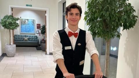 Fernando Odio Esquivel is dressed in a white shirt and black waistcoat. He has a red bowtie. He is pushing a trolley with food on it down a hotel corridor.