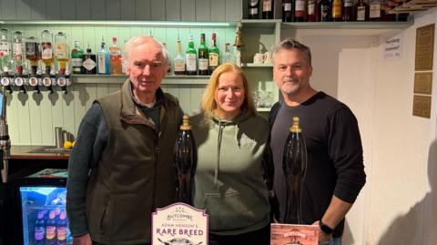 Three people stood behind a bar. There's an older chap on the left and a women in the middle. There's a taller chap on the far right. They are stood behind the pint pulling.