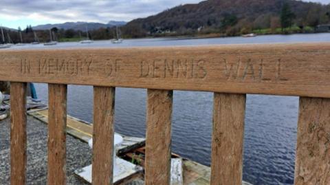 Close up of a wooden bench on the pier of a lake. It has the words 'IN MEMORY OF DENNIS WALL' inscribed along the top. 