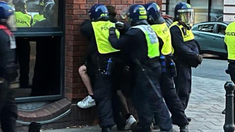 Police officers appearing to restrain someone against a wall in Bristol during the unrest. There are three officers behind the person, whose face cannot be seen, whilst another officer stands nearby looking outwards. They are wearing high-vis jackets and helmets with face shields. 