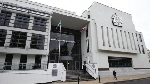 An outside view of Warwick Crown Court, a modern white building with a royal crest and sign reading Warwickshire Justice Centre on the front. A union flag is flying in front of the building.