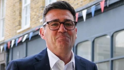 Andy Burnham looks directly forward, his lips are pursed and he wears tortoise shell glasses. His suit jacket is navy and his shirt is white.