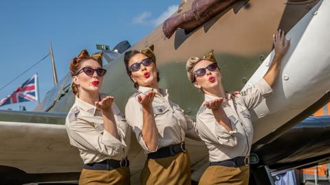 Three women in 1940s-style uniforms and sunglasses pose beside a vintage military aircraft, holding out their hands as if blowing kisses. Each person is wearing a light beige buttoned shirt with sleeves rolled up, dark belts and cat-eye sunglasses. Their hair is styled in vintage pinned-up curls typical of the wartime era.