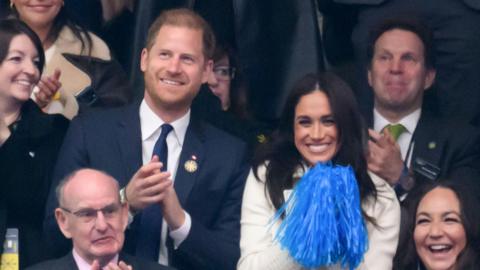 Harry and Meghan grin from the stands. Harry is wearing a suit and Meghan is holding two blue pom-poms