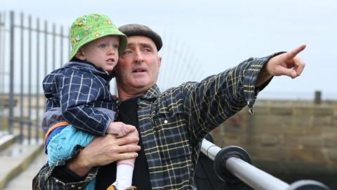 A father carrying his son at the seaside, pointing into the distance.