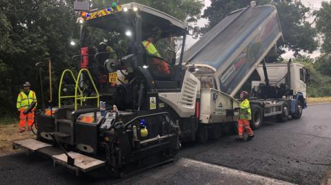 Close-up view of freshly laid asphalt on a rural roadway. A road resurfacing team in high-visibility orange safety clothing operates paving equipment under an overcast sky, with fields stretching alongside the road.