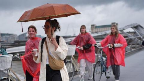 Ladies with bikes and raincoats, and one woman with an orange umbrella, walking over a bridge in the rain. 