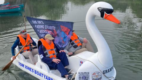 A swan-shaped pedalo with four people on board sets off on the River Thames in Lechlade. The two men at the front are pedalling while the crew members at the back row. There is a large banner between the crew for Combat Stress, a mental health support charity for veterans. All members of the crew are wearing bright orange life jackets. 