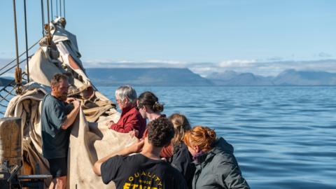 A team of six people lower a traditional tall ship's sail with a calm sea and mountains visible in the background.  