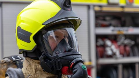A close up shot of a firefighter wearing helmet and breathing apparatus in front of a fire engine