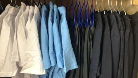 A rail of uniforms with white shirts, blue shirts, grey trousers and black or grey sweatshirts on hangers.