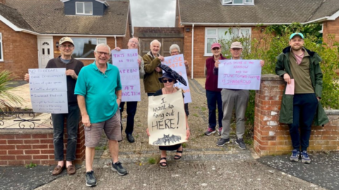 A group of people in front of two bungalows, holding up cardboard signs