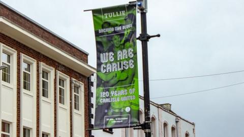 A green banner attached to a black lamppost with a photo of men hugging. The banner reads "Tullie- Backing the Blues- We Are Carlise- 120 years of Carlisle United." Carlisle is spelled with one l in the words 'We are Carlise'.