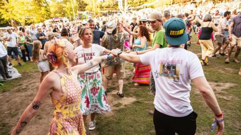 A group of three people with their arms outstretched to the middle of a circle with a large festival crowd in the background