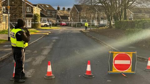 A police officer stands next to some cones on Lea Mill Park Drive. The cones are blocking part of the street off to the public. In the distance, another officer can be seen guarding the other entrance to the scene, which has been marked with police tape.